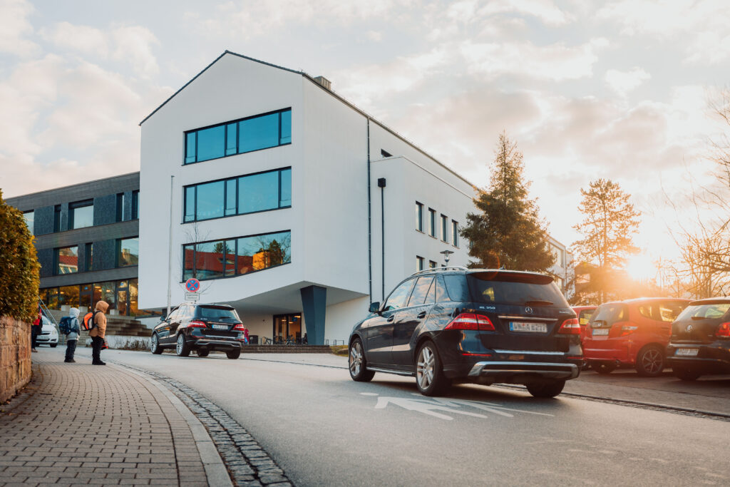 Moderne Gebäude mit großen Fenstern, Autos parken und fahren auf der Straße, Menschen gehen auf dem Gehweg, Sonnenuntergang im Hintergrund.