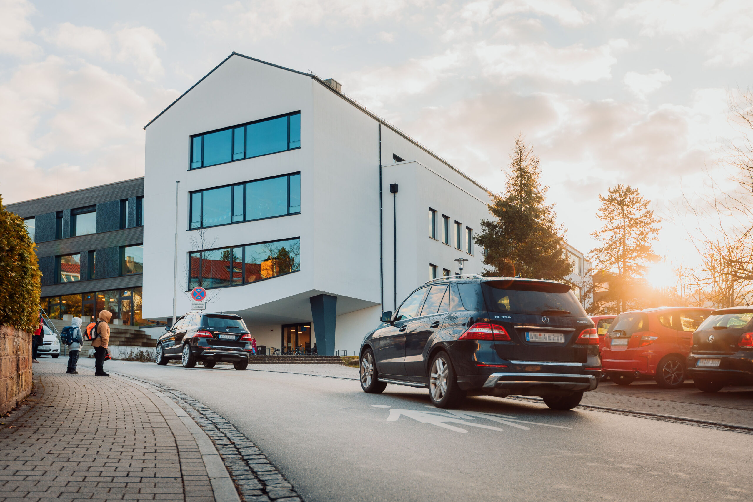 Moderne Gebäude mit großen Fenstern, Autos parken und fahren auf der Straße, Menschen gehen auf dem Gehweg, Sonnenuntergang im Hintergrund.