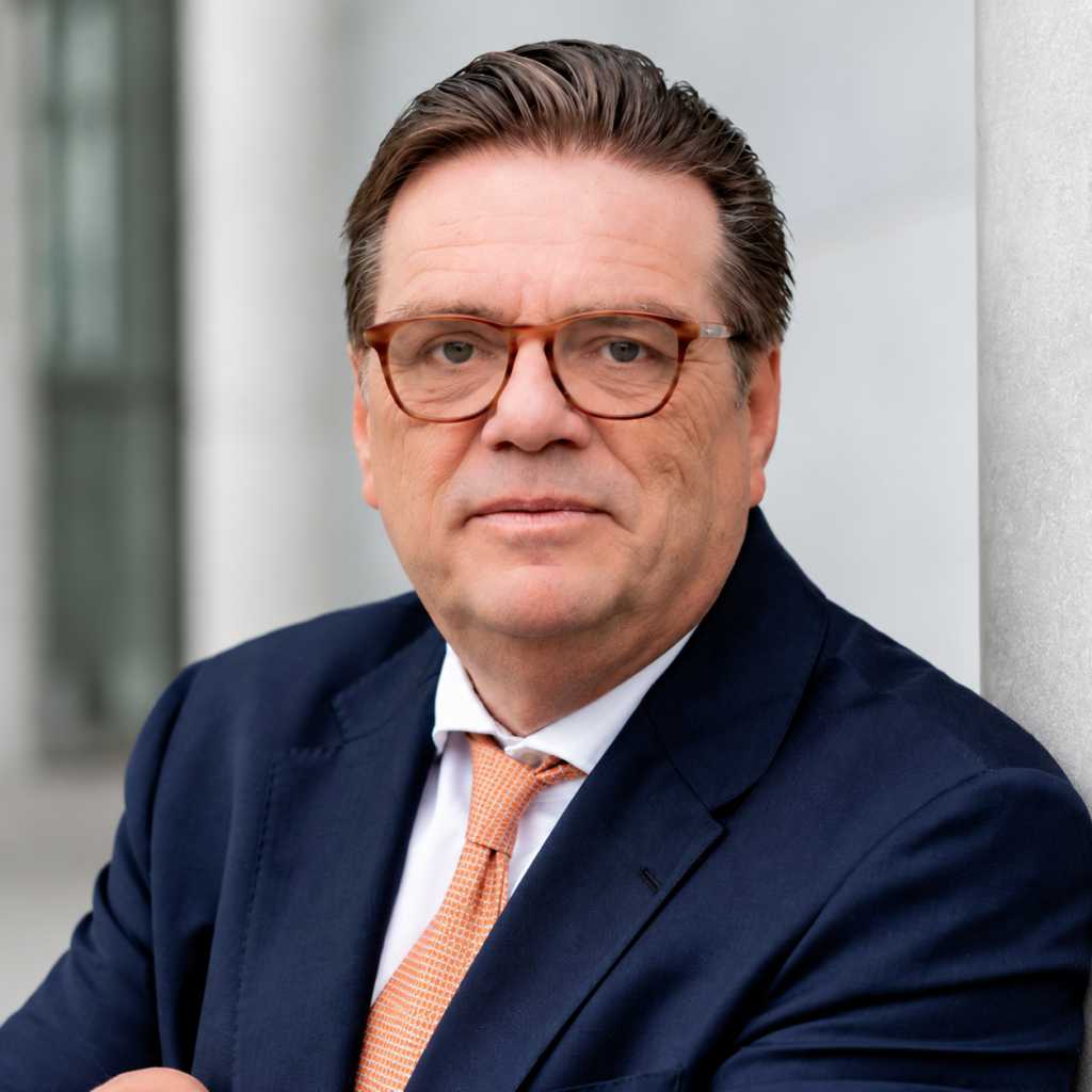 Mittelalterlicher Mann mit Brille und Anzug steht draußen vor einer Wand und schaut direkt in die Kamera.