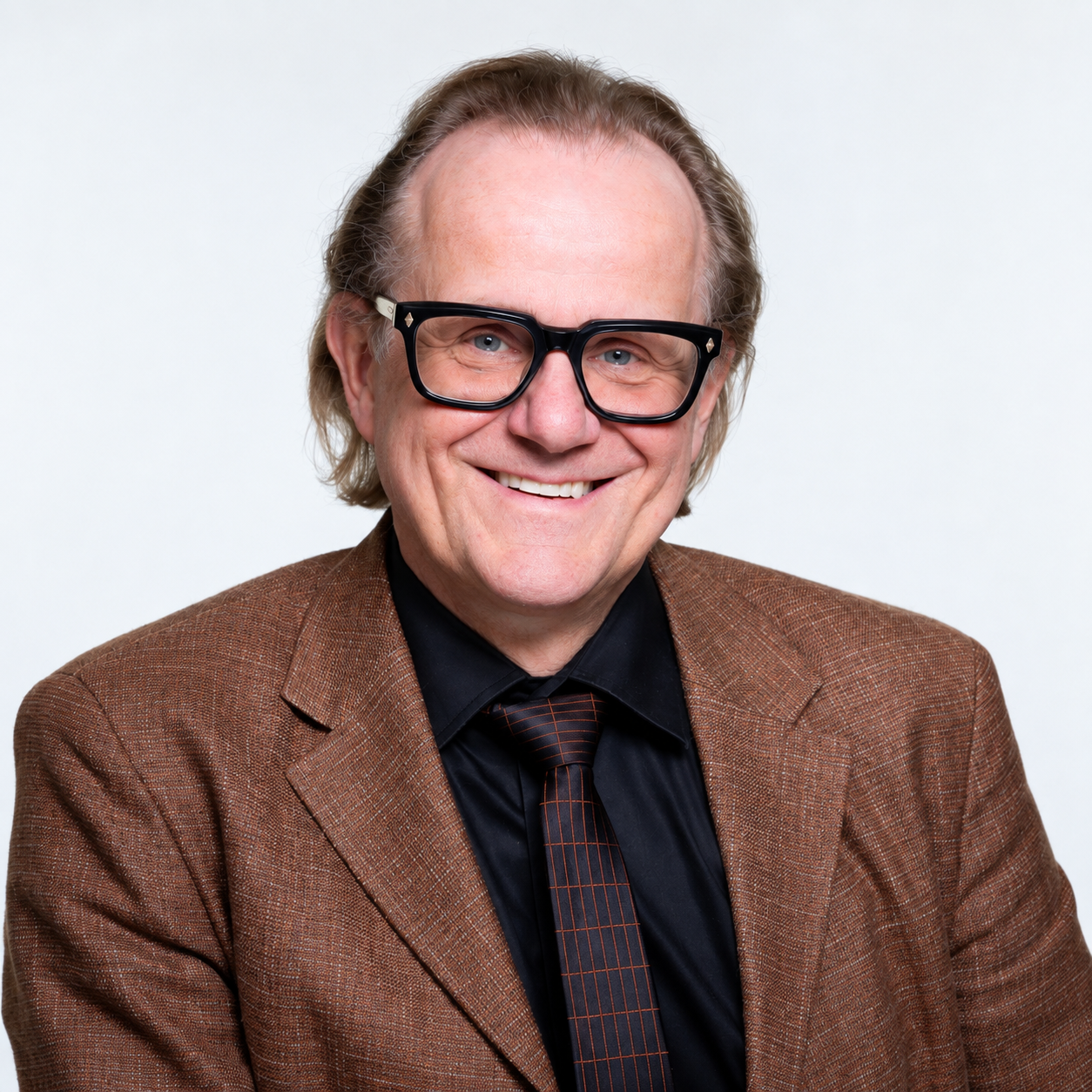 Ein älterer Mann mit Brille, braunem Sakko und schwarzer Krawatte lächelt in die Kamera vor einem hellen Hintergrund.