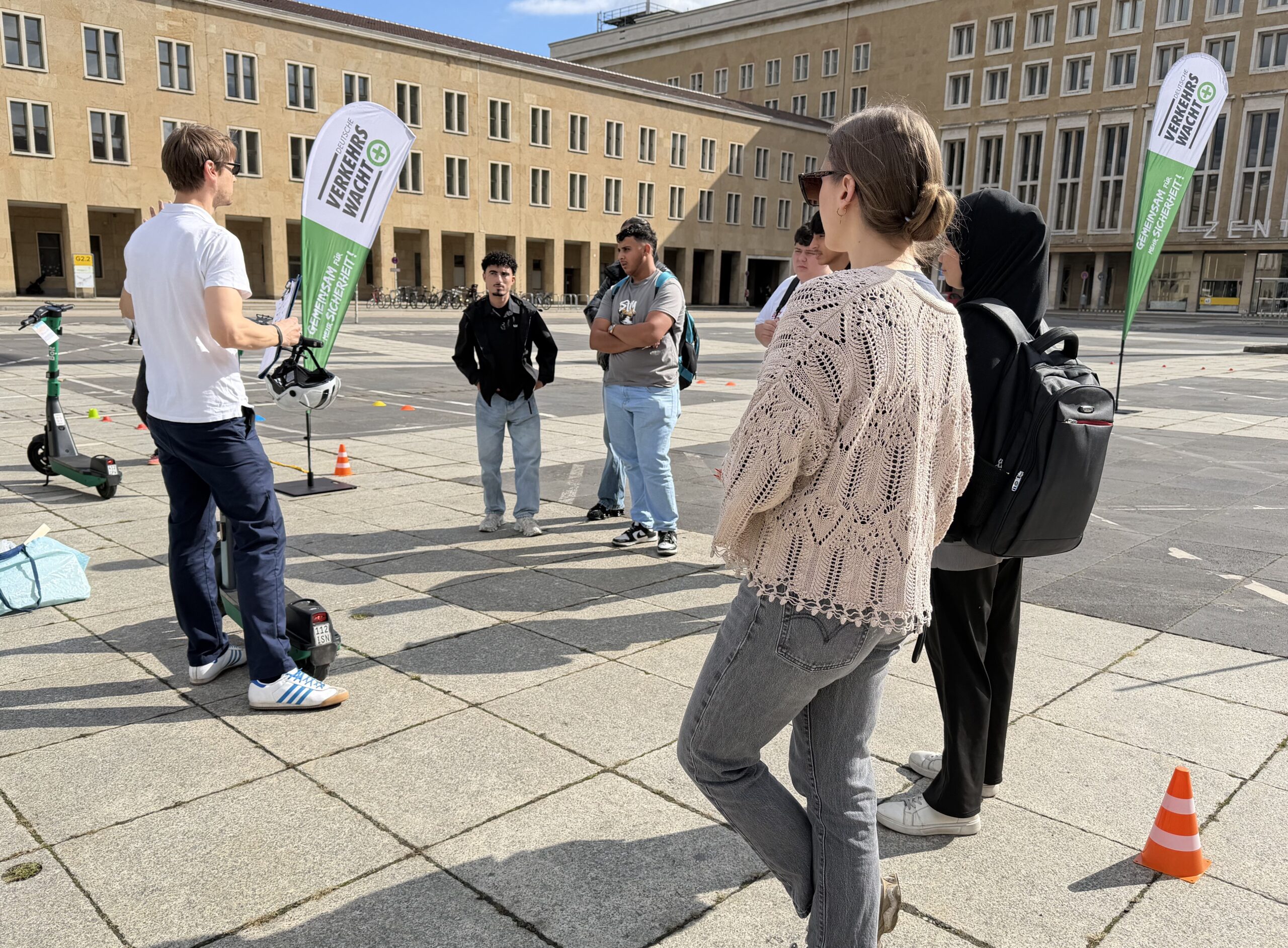 Eine Gruppe junger Menschen hört einem Mann bei einer Verkehrssicherheitsveranstaltung im Freien zu, E-Scooter und Banner sind sichtbar, sonniges Wetter.